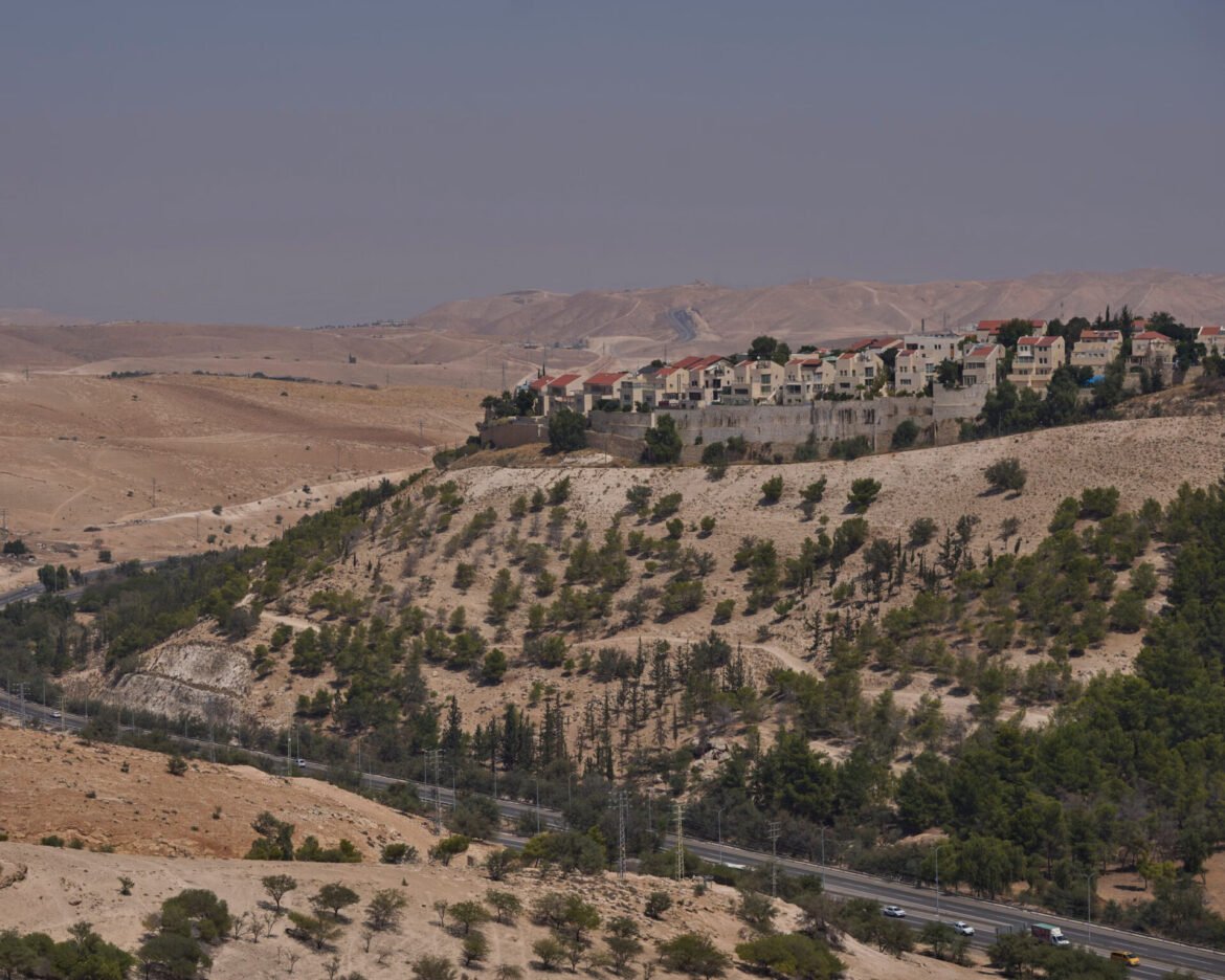 Row of beige houses with red roofs on a hillside retaining wall, set in a dry, arid landscape with a highway in the foreground.