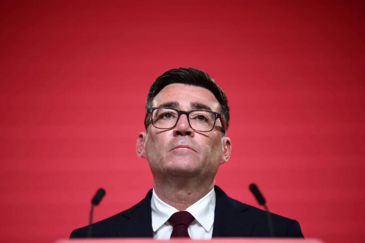 Man in a dark suit and glasses speaking at a podium against a solid red background.