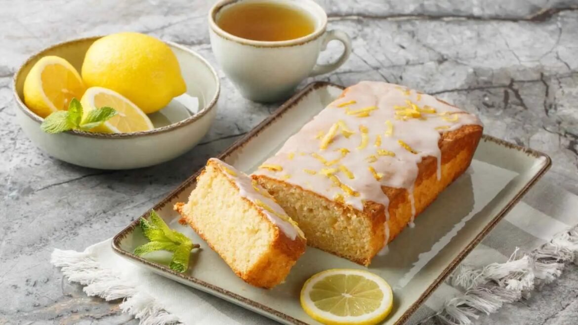 Lemon loaf with white glaze and lemon zest on a rectangular plate, lemon slices and mint nearby, with a cup of tea in the background.