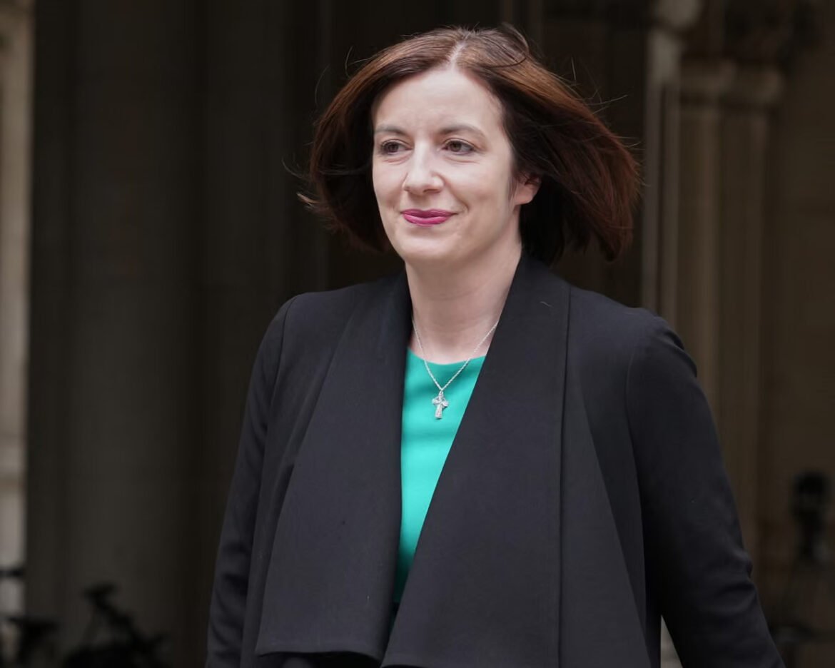 Professional woman walking outdoors, wearing a black blazer over a teal blouse, with a silver cross necklace and a gentle smile.