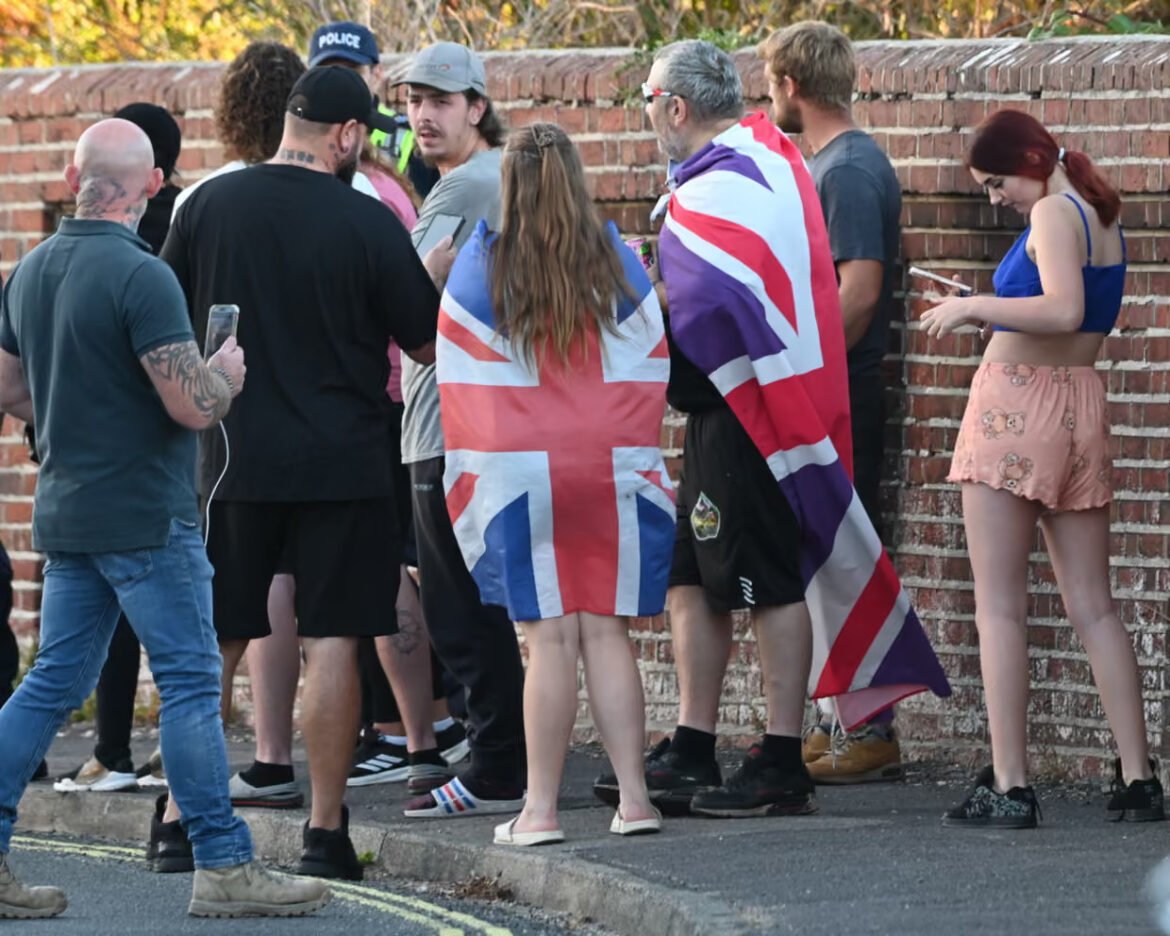 Group of people standing along a brick wall, several wrapped in Union Jack flags, using phones.