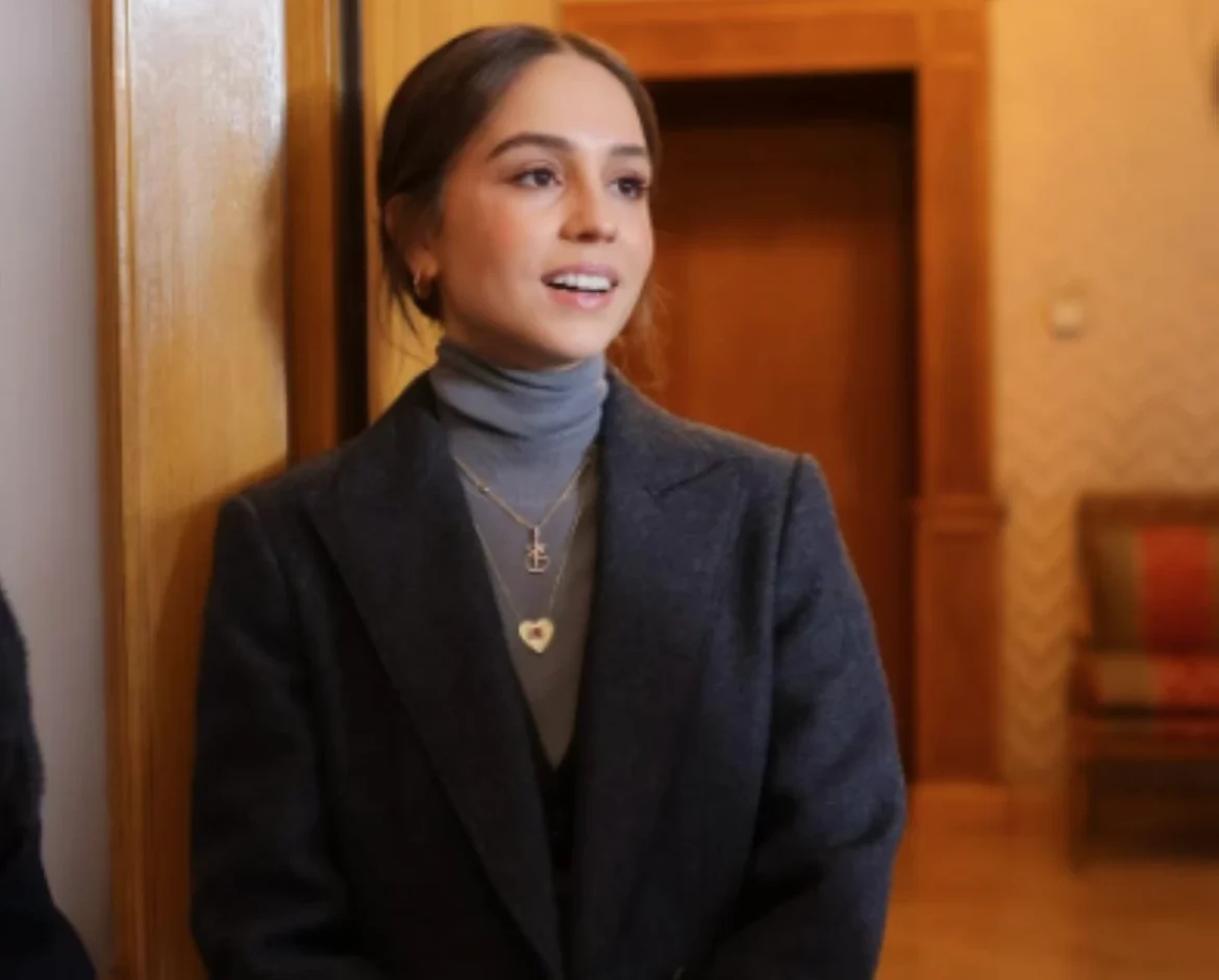 Woman in a gray turtleneck and blazer leaning against a wooden wall, smiling softly.