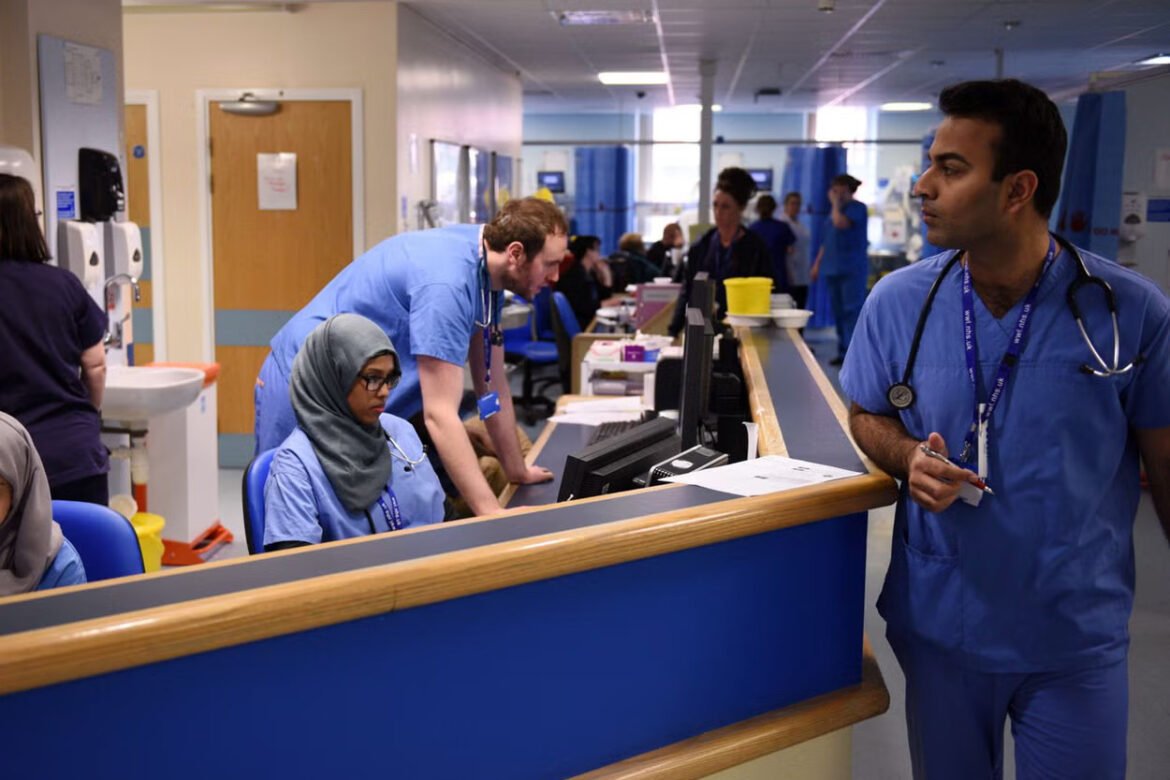 Hospital reception area with healthcare workers at a long counter; a man in blue scrubs stands on the right while a woman wearing a gray hijab sits at a computer station.