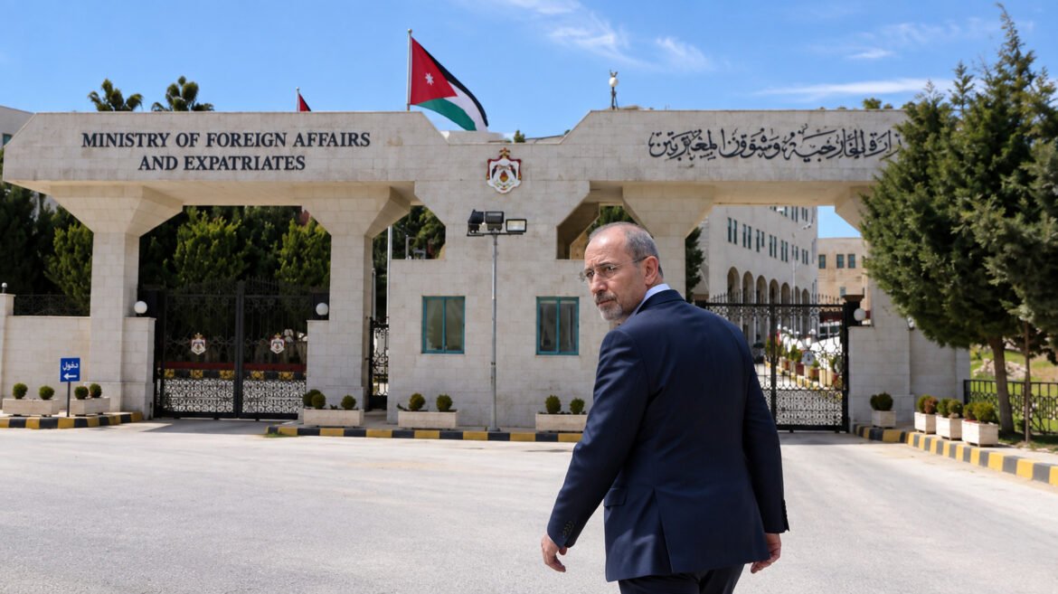 Man in a navy suit walking past the Ministry of Foreign Affairs and Expatriates building, Jordan flag overhead, gates in the background.