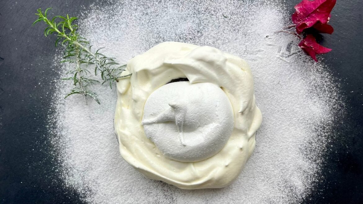 Creamy dessert with a round scoop on top, dusted with sugar, garnished with rosemary and a pink flower on a dark slate surface.