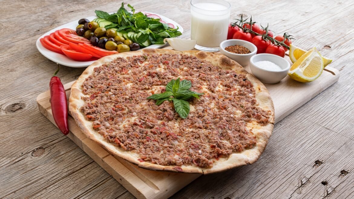 Meat-t topped pizza on a wooden board with a side salad (tomatoes, olives, cucumber, mint) and lemon wedges on the table.