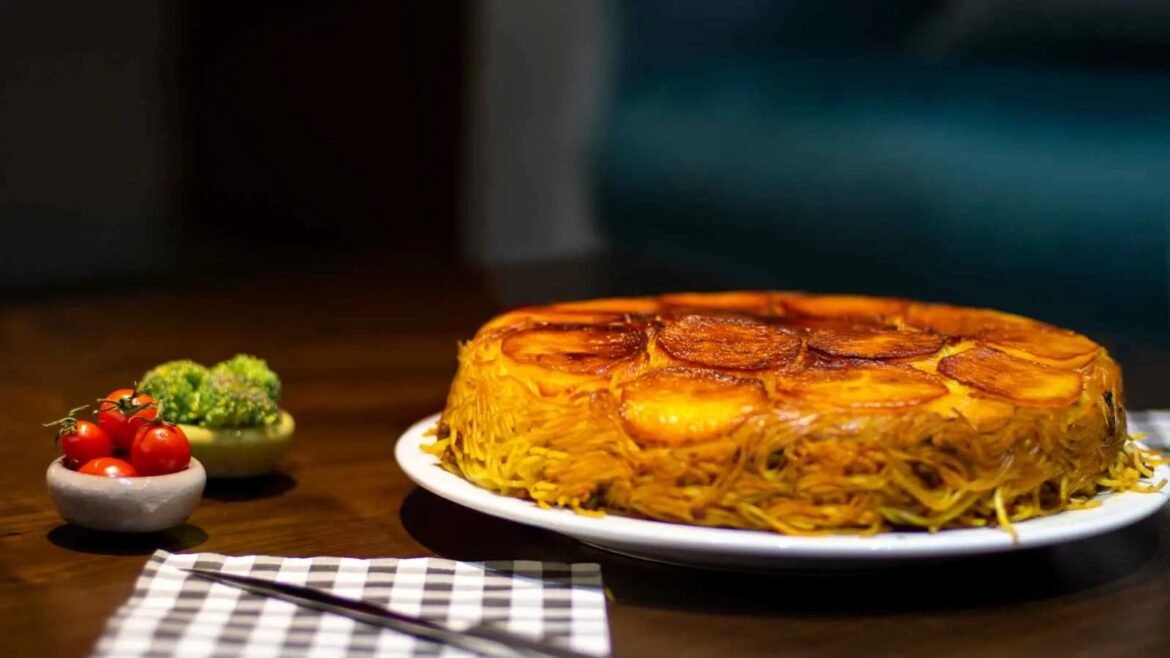 Plate of a crispy noodle cake topped with sliced potatoes, on a white plate with cherry tomatoes and broccoli on the side.