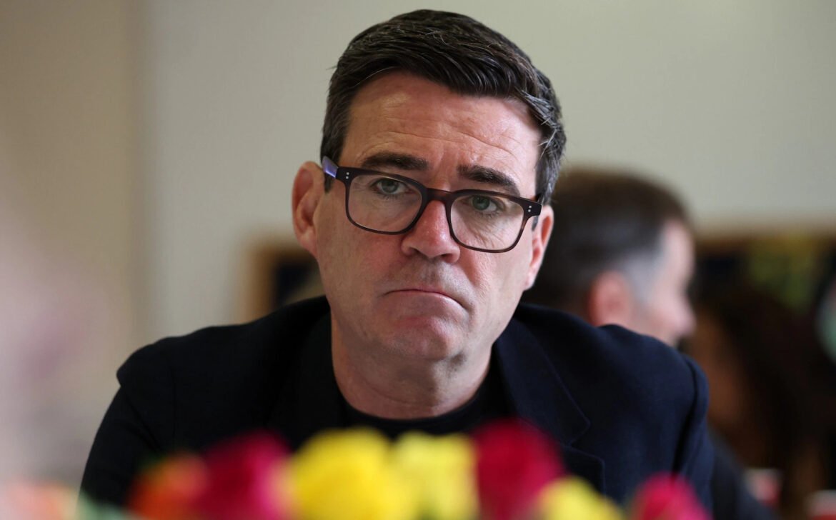 Close-up of a man with dark hair and glasses looking at the camera, indoors, with blurred colorful flowers in the foreground.