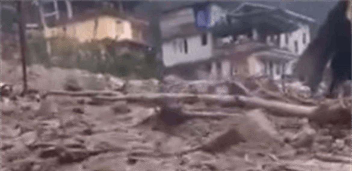Rubble-strewn street with collapsed and damaged houses in the background after a disaster; a dark figure stands on the right among debris