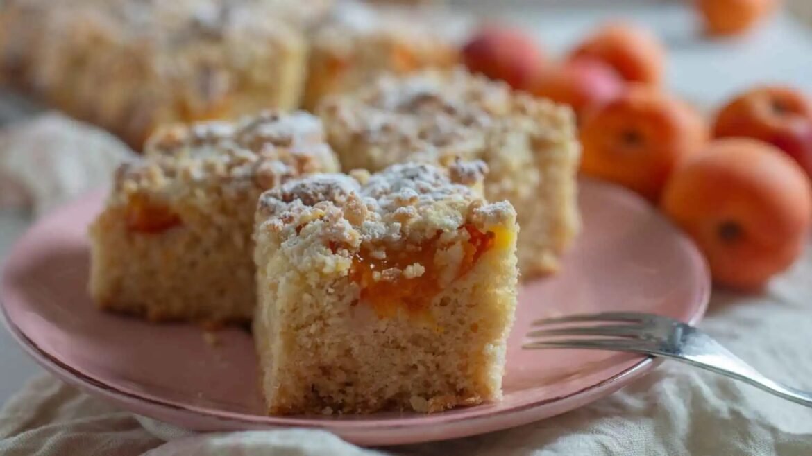 Squares of apricot crumble cake on a pink plate with a fork nearby; soft-focus background with peaches in warm tones.