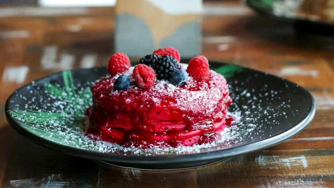 Red berry dessert stacked in a ring on a dark plate, dusted with powdered sugar and topped with raspberries and blueberries.