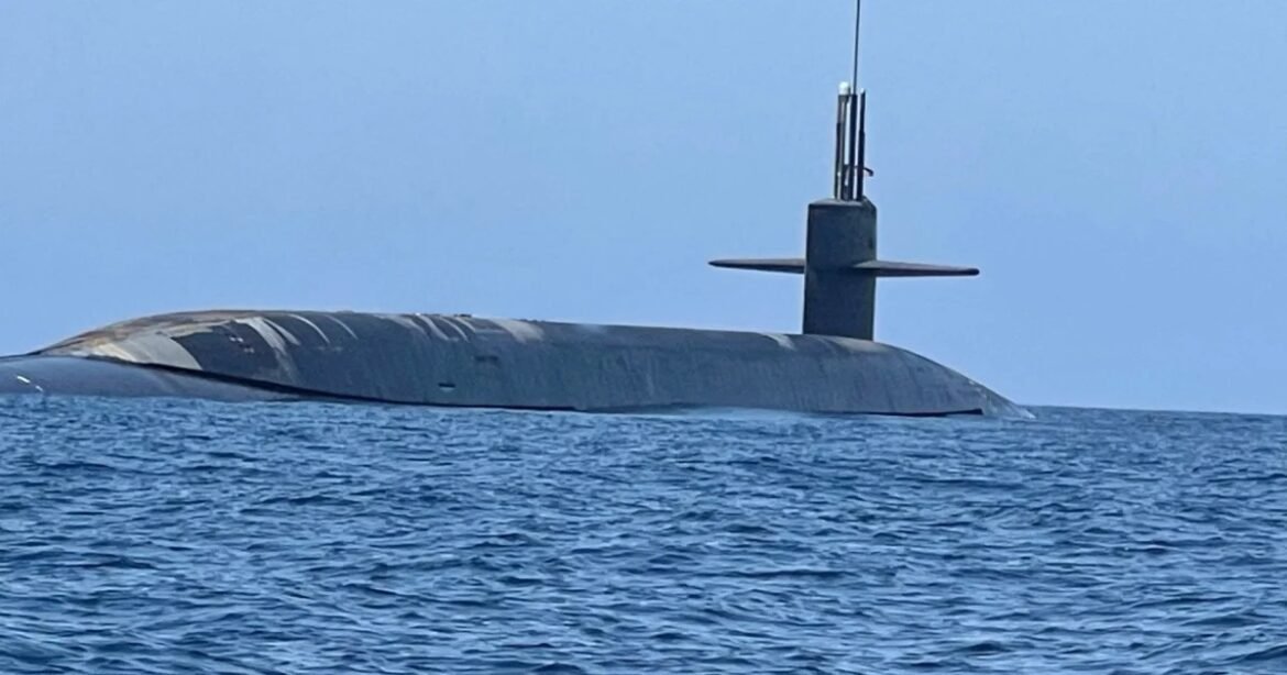 Gray submarine partially surfaced on calm blue ocean, with its sail and mast visible above the water in a clear sky.