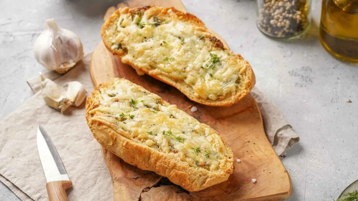 Crusty baguette halves topped with melted cheese and herbs on a wooden board with garlic nearby.