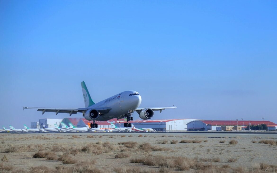 Airliner touching down on a runway at a desert airport, with multiple parked planes and terminal buildings in the background against a clear blue sky.