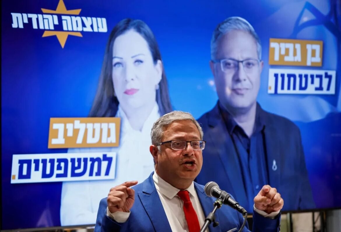 Speaker in a blue suit and red tie addresses microphones in front of a campaign backdrop showing two large portraits and Hebrew text banners behind him.