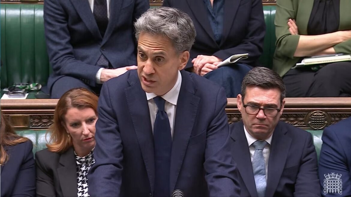 Man in a navy suit speaking at a podium in a parliamentary chamber, with colleagues seated behind him on green benches.
