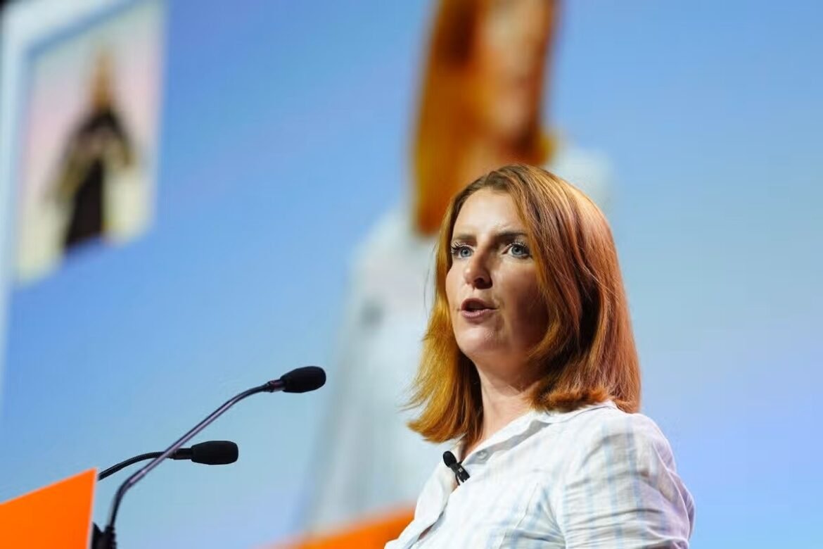 Woman with red hair delivering a speech at a podium with two microphones on a blue backdrop.