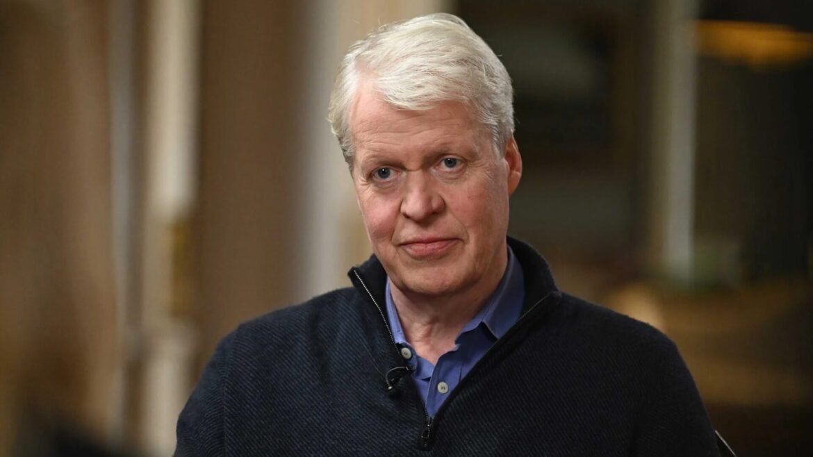 Portrait of an older man with white hair wearing a dark sweater and blue shirt, looking at the camera.