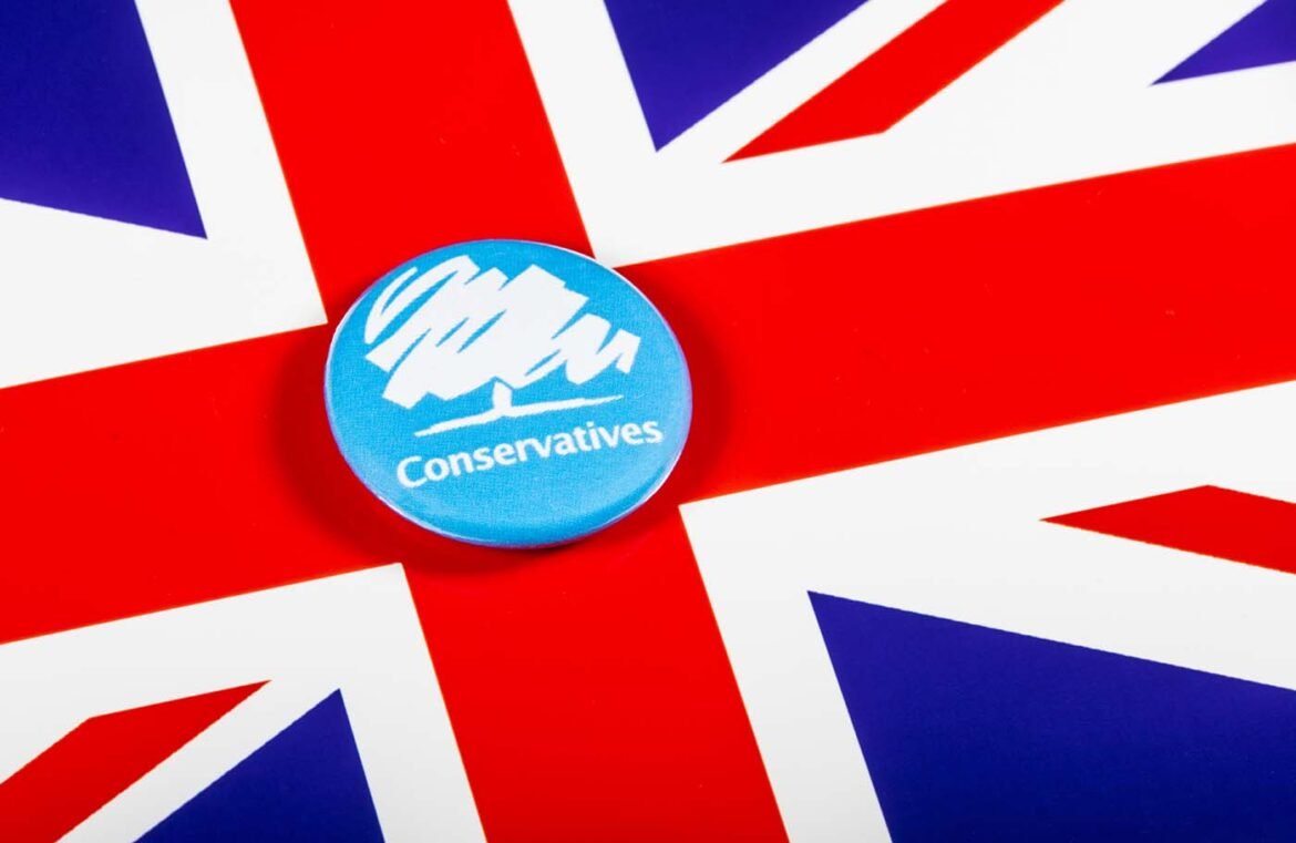 Blue Conservative party badge with a white mountain logo on a Union Jack background showing bold red, white, and blue colors.