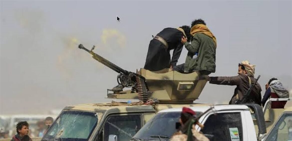 Group of armed men standing on a tan armored vehicle firing a mounted gun in a dusty desert setting.