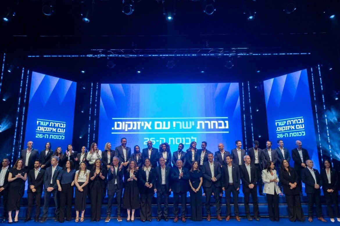 Group of people in formal attire standing on a blue-lit stage for a ceremony, with Hebrew text on screens behind them.