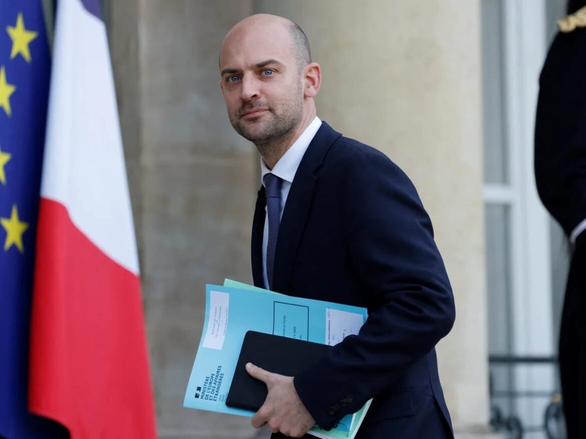 Man in a dark suit carrying blue folders and a black notebook outside a government building with flags in the background.