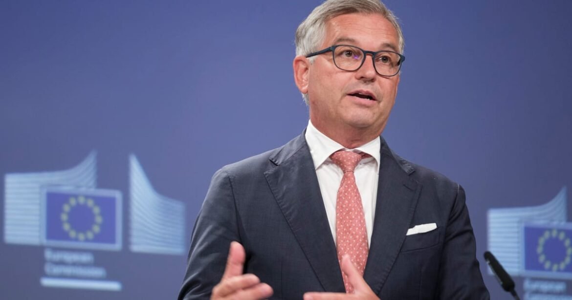 Man in a dark suit, pink tie, and glasses speaks at a press conference with EU graphics in the background.