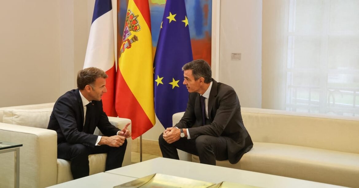 Two men in dark suits talk while seated on white couches, with France, Spain, and EU flags behind them in a formal setting