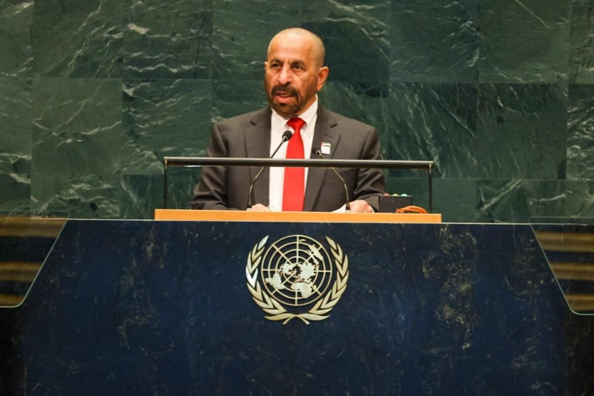 Man in a dark suit and red tie stands at a UN podium delivering a speech, with the UN emblem on the front panel behind him.
