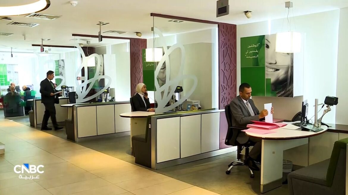 A modern bank office with tellers at curved counters, customers at desks, and a man reviewing papers at a rounded frontline desk; CNBC Arabia logo in bottom-left.