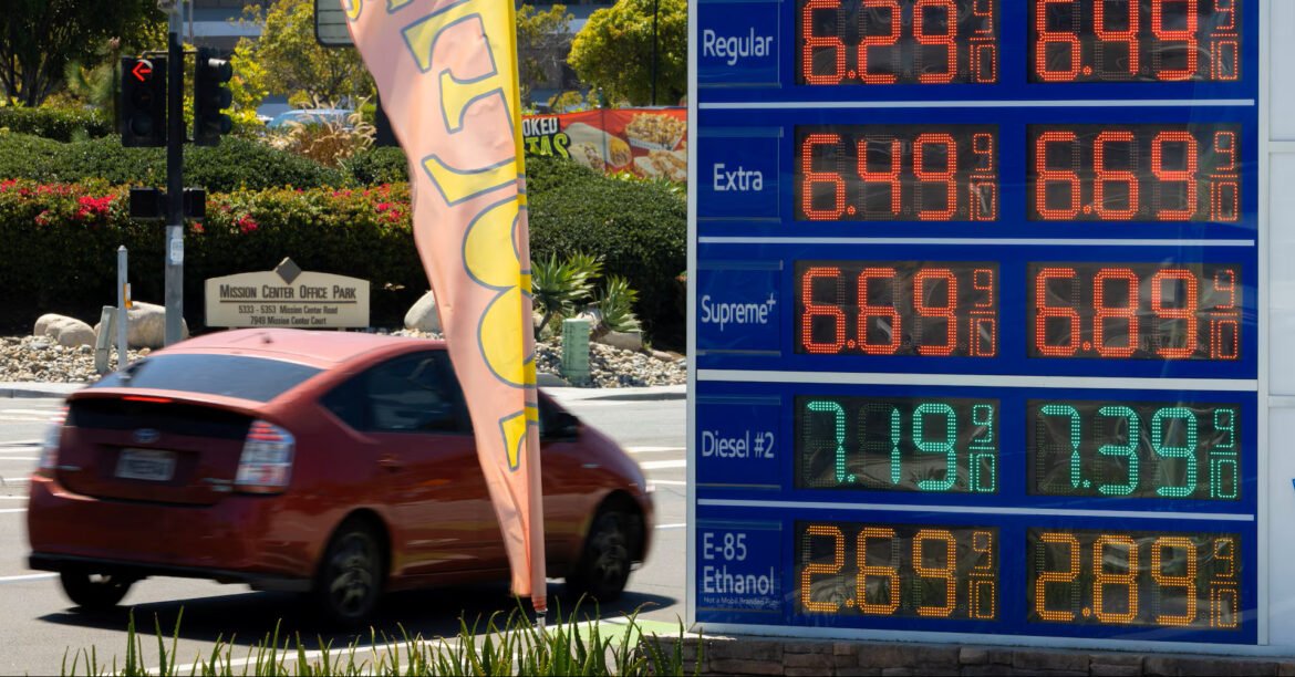 Large blue fuel-price board with orange LED prices beside a red car on a street.
