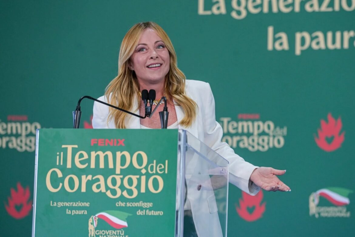 Smiling blonde woman in a white blazer speaks at a green podium with microphones at a political event.