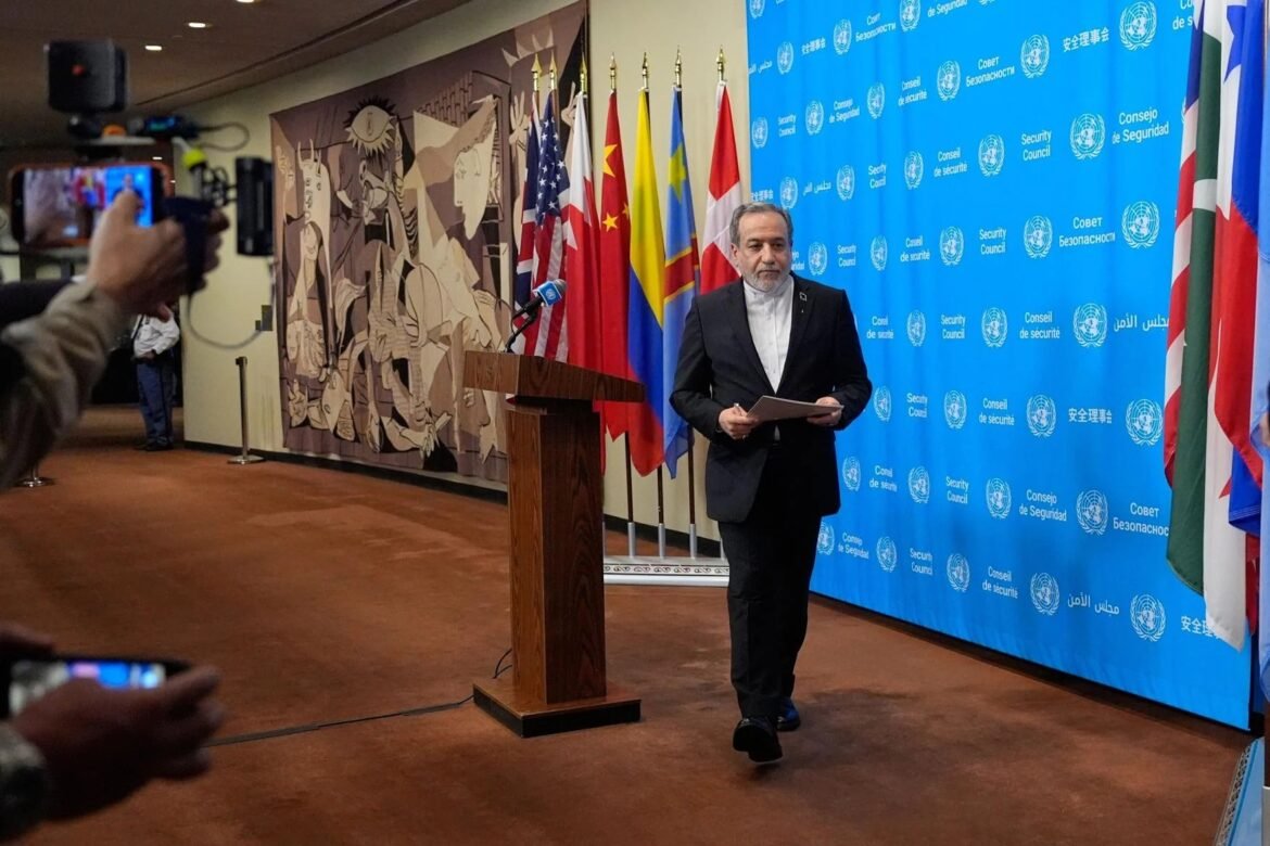 Man in a dark suit stands at a wooden podium with papers, in front of a blue United Nations Security Council backdrop and flags. A photographer films him from the left.