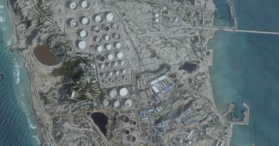 Aerial view of an industrial facility with many circular storage tanks along a coastline, including a harbor and breakwater to the right.
