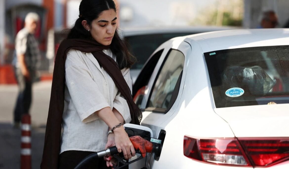 A woman refueling a white car with a red fuel nozzle on a city street, looking down at the nozzle.