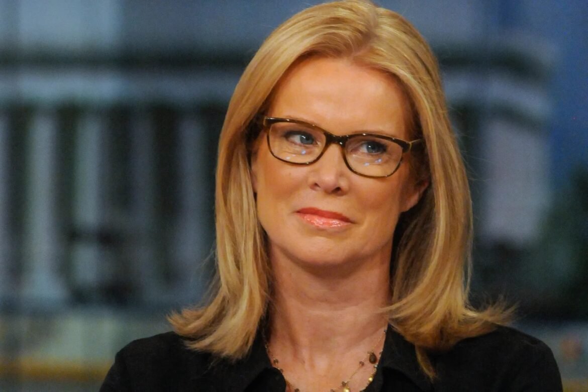 Blonde woman wearing glasses and a black blouse, looking to the right with a slight smile.