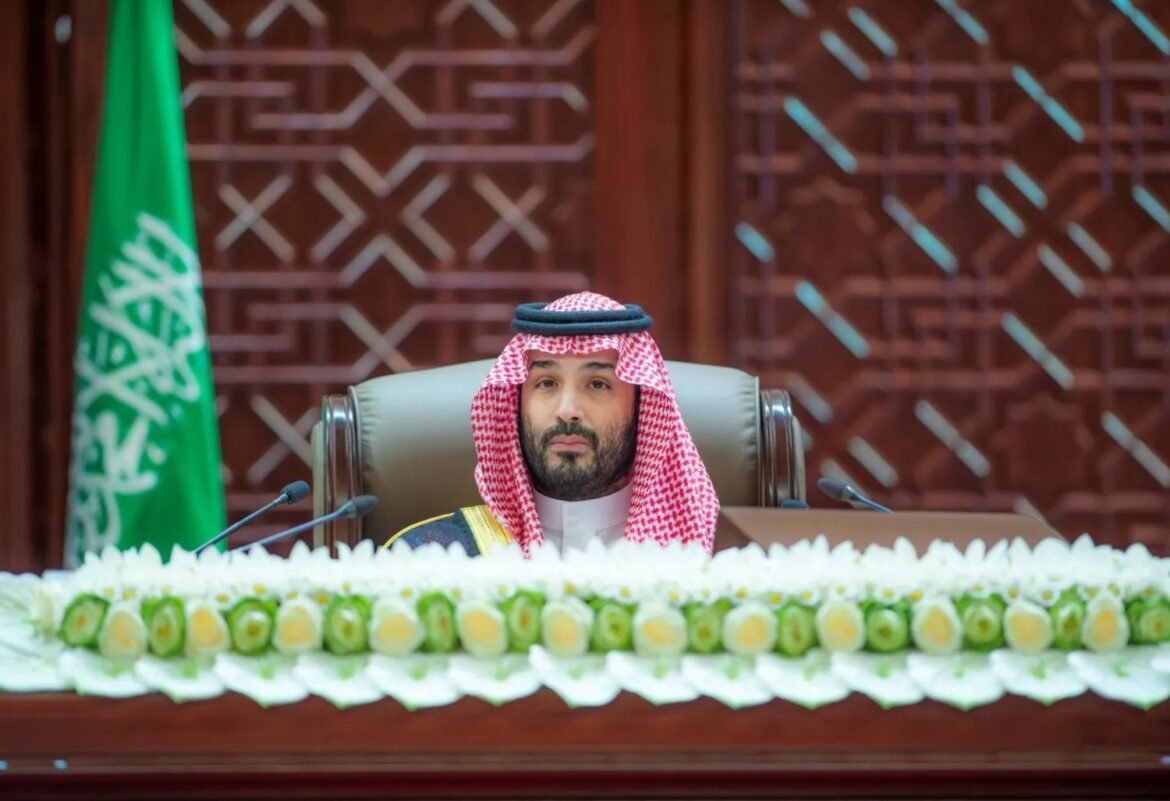 Man in traditional Saudi attire sits at a dais with a floral garland and microphones, green Saudi flag in the background.