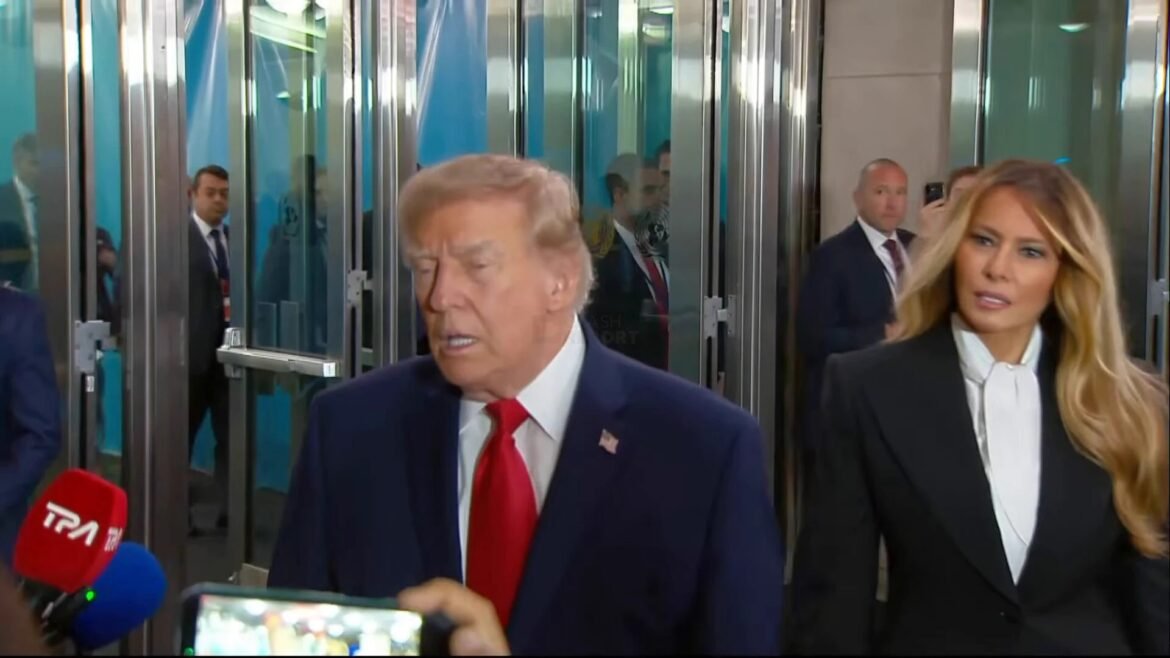 Man in a dark suit and red tie speaks to reporters outside glass doors; a woman in a blazer stands nearby.
