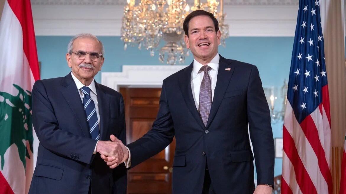 Two men in suits shake hands in a formal room, with a chandelier and flags (Lebanese on the left, U.S. on the right) in the background.