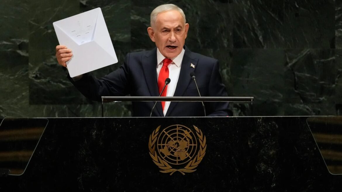 Man in a dark suit and red tie speaks at a United Nations podium, raising a large white folder with papers above his head.
