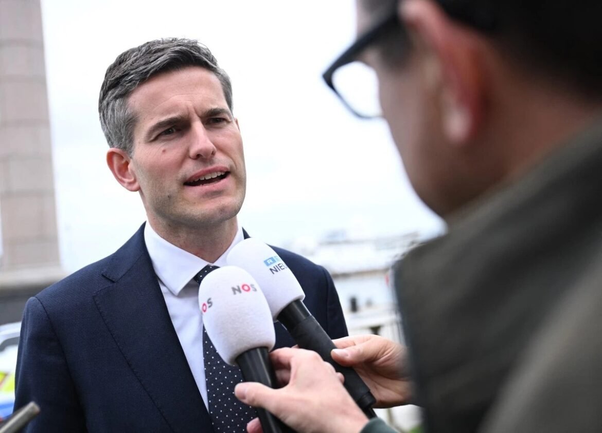Man in a navy suit being interviewed outdoors, speaking into two microphones held by reporters in front of him.