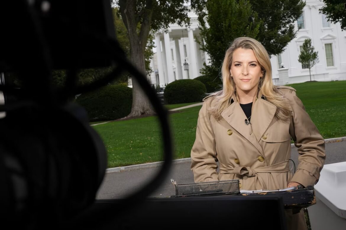 Female reporter in a beige trench coat speaks outdoors, microphone clipped to her lapel, in front of a white neoclassical building and green lawn.