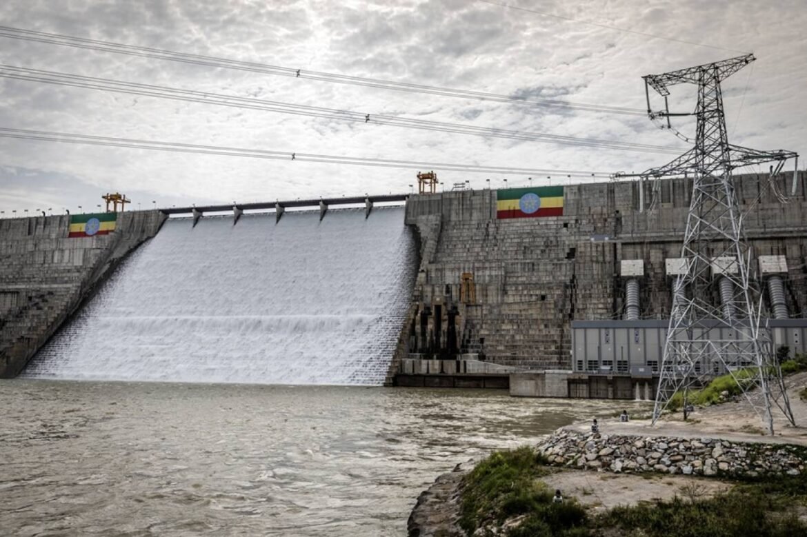 Large concrete dam with water spilling over the spillway and power lines crossing the sky in the background.