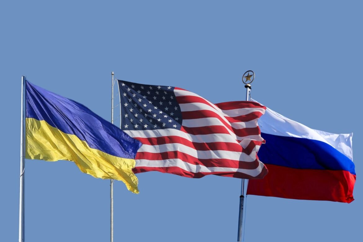 Three national flags (Ukraine, United States, and Russia) waving in the wind against a clear blue sky.