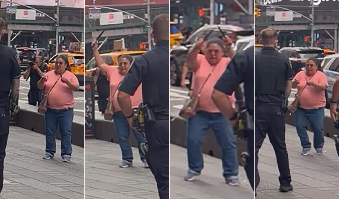Woman in pink shirt with crossbody bag on a busy city sidewalk, facing an armed security officer; photographers and yellow taxis in the background.