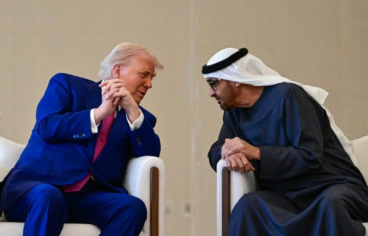 Two men sit facing each other, leaning in for a private conversation; one in a blue suit, the other in traditional Middle Eastern robes and a head scarf.