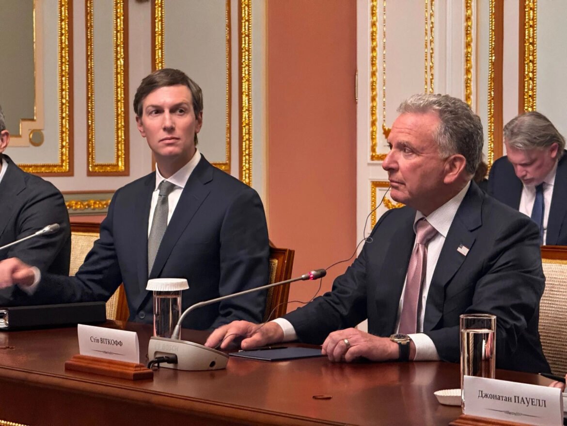 Two men in dark suits sit at a wooden conference table with microphones in a gilded, ornate room during a formal meeting.