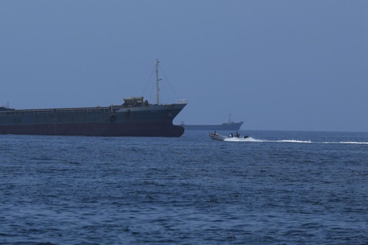 Two ships on the open sea: a massive cargo freighter on the left and a small speedboat creating a white wake to the right.