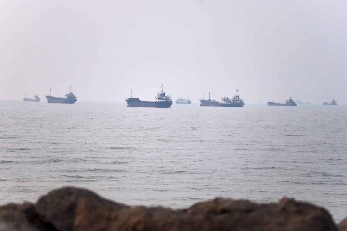 Flock of cargo ships on a calm, hazy sea seen from a rocky shore in the foreground.