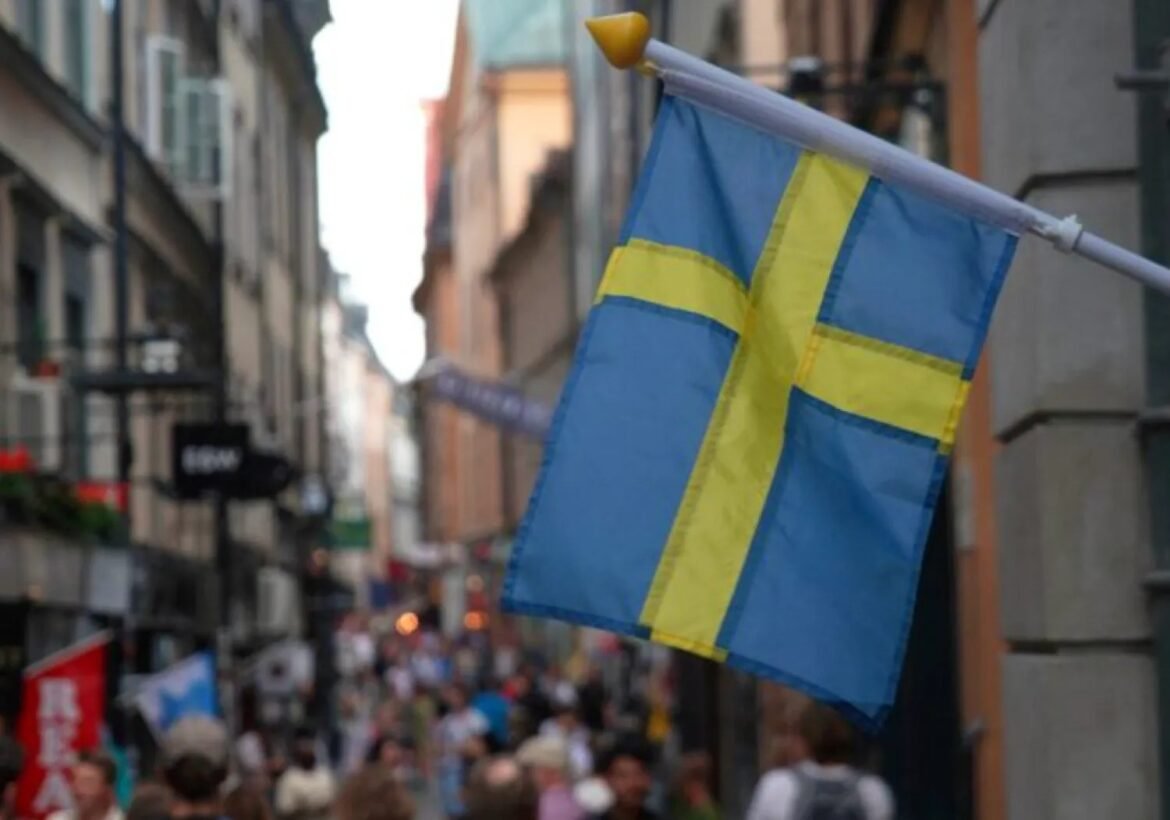 Blue and yellow Swedish flag on a pole over a busy urban street with blurred pedestrians in the background, suggesting a street festival or city scene.
